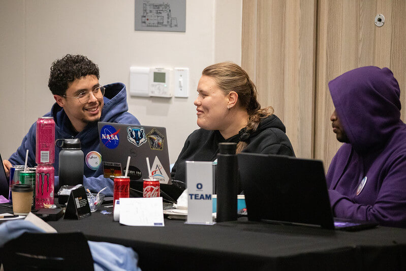 Three hackathon participants sit at a table with laptops open, collaborating and discussing solutions.